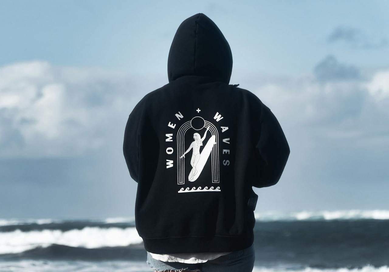 A backshot of a woman in a blue “Women + waves” hoodie standing in front of an ocean.